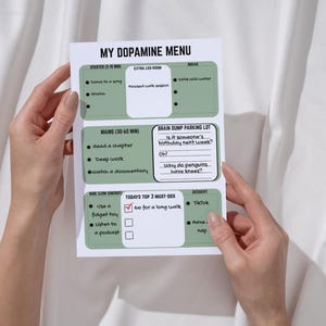 Könnte beinhalten: Ein weißes Papier mit dem Titel "MY DOPAMINE MENU" mit grünen Abschnitten, in denen Aktivitäten wie "dance to a song" und "go for a long walk" aufgeführt sind. Das Papier wird von zwei Händen gehalten.