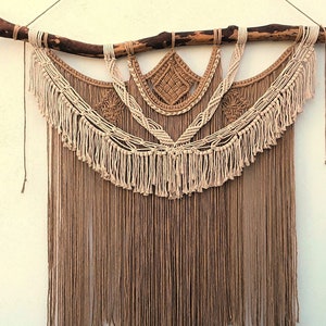 May include: A large macrame wall hanging featuring a natural wooden branch at the top. The design incorporates beige and brown knotted cords, creating a textured, geometric pattern with a fringed bottom. The wall decor is suspended by a thin, natural-colored cord.