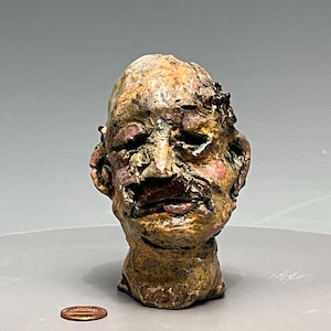 May include: A textured, hand-sculpted ceramic head sculpture featuring a mottled brown, tan, and pink glaze with deep crevices and uneven facial features. A copper-colored US penny is placed next to the sculpture for scale reference.