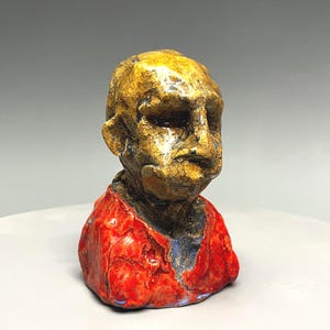 May include: A ceramic bust sculpture featuring a head with a textured, crackled yellow and brown glaze finish. The figure wears a vibrant, glossy red garment with a visible blue-toned underlayer, showcasing a rough, hand-molded clay texture.
