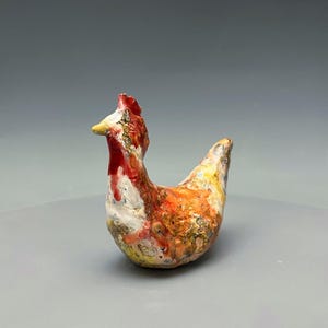 May include: A small ceramic chicken figurine featuring a textured surface with a mottled color palette of red, orange, yellow, and white. The sculpture depicts a standing chicken with a prominent red comb, a yellow beak, and a pointed tail.