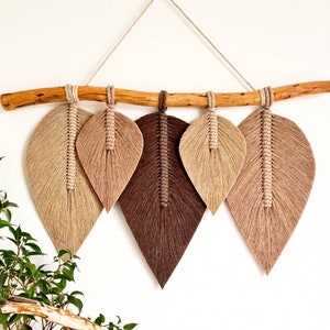 May include: A wall hanging made of macrame with four brown and beige leaves hanging from a natural branch. The leaves are made of knotted string and are arranged in a staggered pattern.