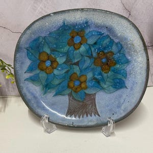 Peut inclure: Une assiette en céramique ronde représentant un arbre peint à la main avec un tronc brun et des feuilles bleu vif. L'arbre est orné de quatre fleurs brunes proéminentes, chacune centrée par un point bleu vif. Le fond est bleu clair moucheté.