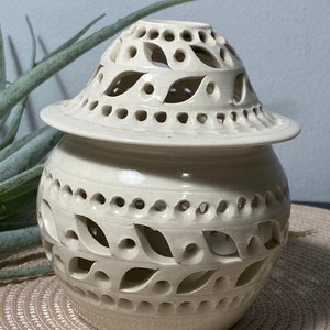 May include: Off-white ceramic jar with a lid. The jar has a round body with leaf-shaped cutouts and a matching lid with similar cutouts. The jar is sitting on a tan surface.