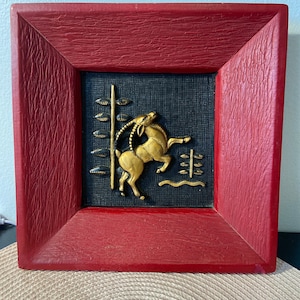May include: A framed wall art piece with a red wooden frame and a black background. The image is of a gold-colored animal, possibly a goat, leaping in a forest setting.