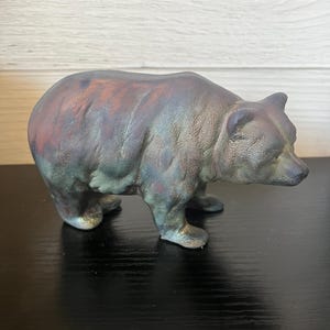 May include: A small, dark green and brown ceramic bear figurine. The bear is standing on all fours and has a slightly tilted head.
