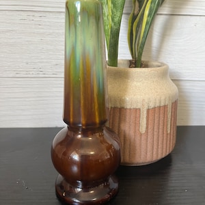 May include: A small, brown and green ceramic vase with a bulbous base and a tall, slender neck. The vase has a glossy finish and a unique, marbled pattern.
