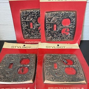 Peut inclure: Quatre plaques murales vintage avec des motifs ornés en finition laiton. Les plaques sont emballées dans un emballage en carton rouge et blanc avec le texte "Stylset Wallplate" imprimé dessus. L'emballage comprend également le texte "Dexter Lock, division of Kysor Industrial Corp. 1601 Madison Ave. S. E., Grand Rapids, Mich. 49502".