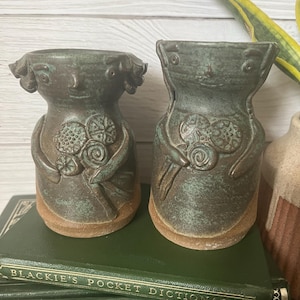 Puede incluir: Dos jarrones de cerámica hechos a mano con un diseño de rostro único. Los jarrones son de color verde y marrón moteado, con una superficie texturizada. Cada jarrón presenta un rostro estilizado y adornos florales. Se encuentran sobre una pila de libros verdes.