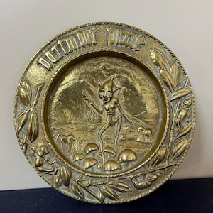 May include: A brass wall plaque with a raised design of a goblin-like figure standing in a forest setting. The plaque is circular and has the words "door knocker piece" inscribed around the edge.