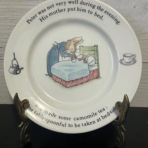 May include: A white ceramic plate with a picture of Peter Rabbit in bed with a cup of tea. The plate has a black border and the text "Peter was not very well during the evening. His mother put him to bed. and made some camomile tea: One table-spoonful to be taken at bed-tim" around the edge.