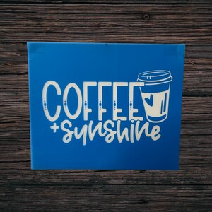 May include: A blue sign with the words "COFFEE + sunshine" in white script and a coffee cup graphic. The sign is rectangular and appears to be made of a solid material. The background is a dark wood surface.