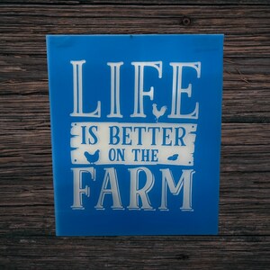 May include: A blue sign with white lettering that reads "LIFE IS BETTER ON THE FARM." The sign features illustrations of a rooster, a hen, and a small leaf. The sign is set against a wooden background.