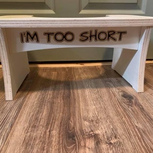 May include: A small, wooden step stool with a white painted frame and a natural wood top. The text "I'M TOO SHORT" is cut out of the wood and adhered to the front of the stool. The stool is designed to help people reach higher places.