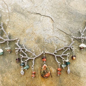 Puede incluir: Un collar de plata con un diseño de rama y piedras colgantes. Las piedras son verdes, rojas y marrones. El collar está sobre un fondo gris.