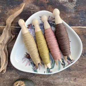 May include: Four spools of thread in varying colors: tan, olive green, dusty rose, and brown. The spools are on a floral patterned dish. A piece of driftwood and a stone with a carved design are also present.