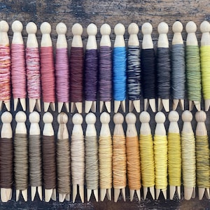 May include: A collection of wooden spools wrapped with colorful threads. The threads vary in shades of pink, purple, blue, black, gray, green, and yellow. The spools are arranged in neat rows on a wooden surface, showcasing a variety of colors.