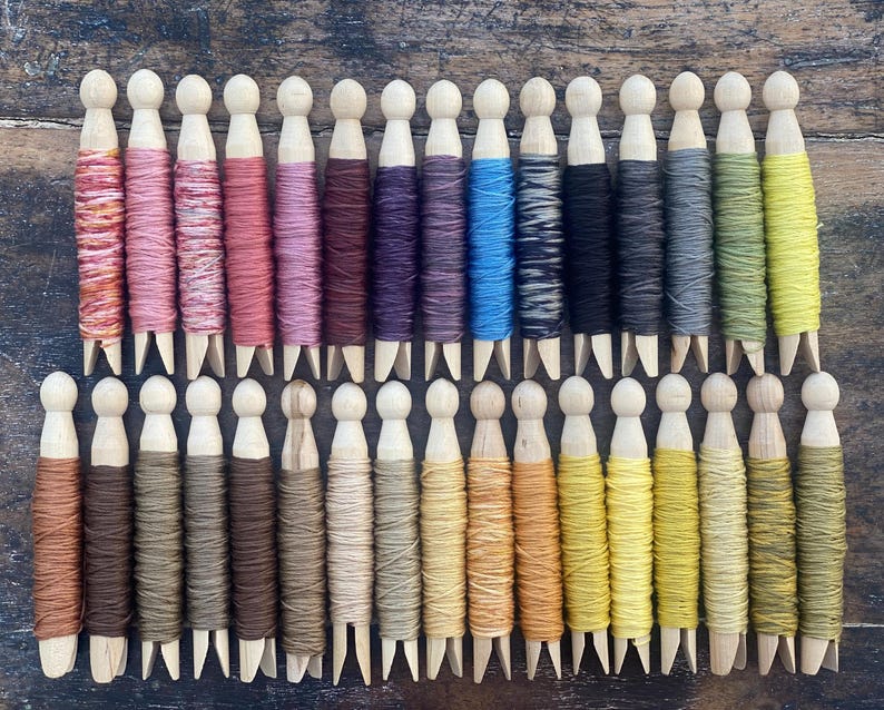 May include: A collection of wooden spools wrapped with colorful threads. The threads vary in shades of pink, purple, blue, black, gray, green, and yellow. The spools are arranged in neat rows on a wooden surface, showcasing a variety of colors.