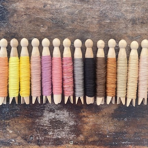 May include: A collection of wooden doll-shaped spools, each wrapped with colorful thread. The threads vary in shades of red, orange, yellow, pink, gray, black, brown, and green. The spools are arranged in a row on a wooden surface.