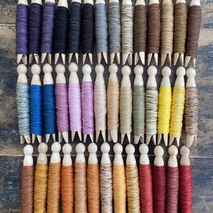 Pu&ograve; includere: Una collezione di mollette da bucato in legno avvolte con filo da ricamo di vari colori, disposte in tre file orizzontali. I fili presentano un ampio spettro di tonalit&agrave; tra cui viola, blu, rosa, giallo, marrone e rosso.