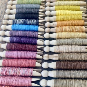 May include: Rows of colorful embroidery floss wound on wooden spools. The floss colors include shades of yellow, gray, black, blue, purple, pink, and brown. The wooden spools have a rounded top and a pointed end.