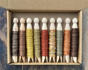 May include: Nine wooden clothespins arranged in a cardboard box, each wrapped with embroidery floss in various earth tones including shades of brown, mustard yellow, olive green, rust orange, and a variegated blend of yellow and green threads.