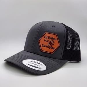 May include: A gray and black trucker hat with a brown leather patch that says "I'd Rather bee Beekeeping" with a honeycomb design. The hat is a snapback style.