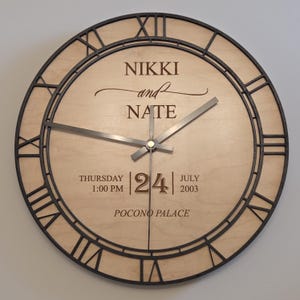 May include: A round wooden clock with a black frame and Roman numerals. The clock face is light-colored with the names "NIKKI and NATE" and the date "24 July 2003". The time is 1:00 PM. The words "POCONO PALACE" are at the bottom.