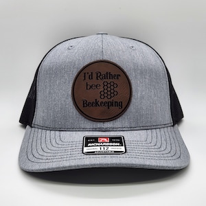 May include: A gray and black trucker hat with a brown leather patch that says "I'd Rather bee Beekeeping". The patch has a honeycomb design. The hat is adjustable and has the Richardson 112 logo on the back.