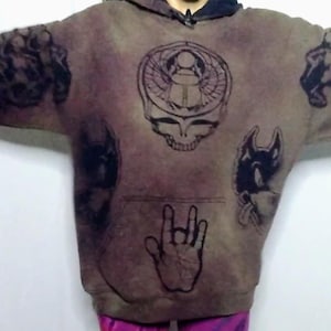 May include: Brown hoodie with multiple black graphic designs, including a skull, a hand gesture, and other illustrations. The hoodie has a front pocket and a hood.
