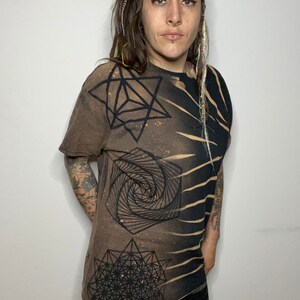 May include: A brown t-shirt with a black geometric design. The design features a spiral, a star, and a flower of life pattern.