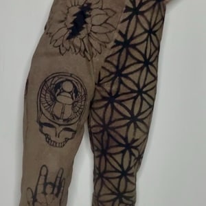 May include: Brown pants with a variety of black and white graphic designs, including a sunflower, a skull, a hand, and a geometric pattern.