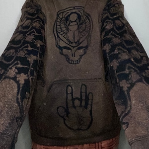 Puede incluir: Sudadera con capucha marrón con un estampado gráfico negro de una calavera con un escarabajo egipcio en su interior. Las mangas tienen un estampado floral negro. La parte delantera de la sudadera también tiene un estampado gráfico negro de una mano haciendo el gesto de rock and roll.