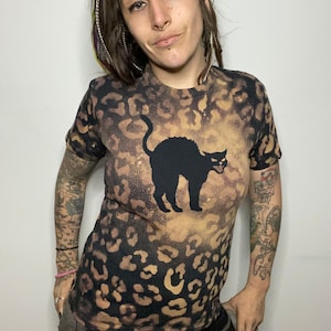 Puede incluir: Camiseta con estampado de leopardo negro y marrón con una silueta de gato negro. La camiseta tiene un efecto decolorado, creando un patrón único. El gato está en una pose temática de Halloween.