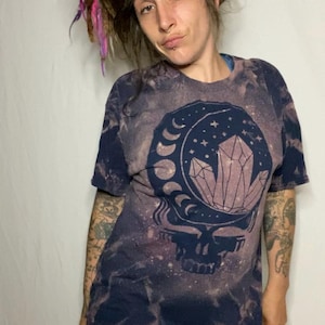 May include: A dark blue tie-dye t-shirt with a bleached design of a skull with a crescent moon and crystals. The shirt has a distressed look.