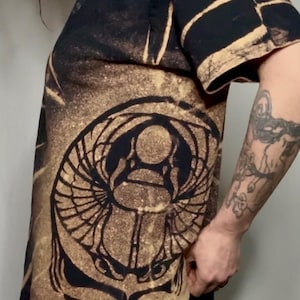 Peut inclure: T-shirt noir avec un motif délavé et vieilli. Le t-shirt présente un grand motif d'un scarabée et d'un crâne. Le design est dans des tons de marron et de noir, créant une esthétique vintage et grunge.