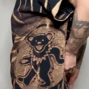 May include: Black and tan tie-dye t-shirt featuring a dancing bear graphic and a skull design. The shirt has a crew neck and short sleeves. The bear graphic is in black and the skull is at the bottom of the shirt.