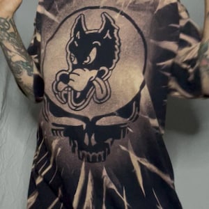 May include: A tie-dye t-shirt featuring a black wolf graphic within a circle, above a skull design. The shirt has a brown and black color scheme. The design is reminiscent of the Grateful Dead band.