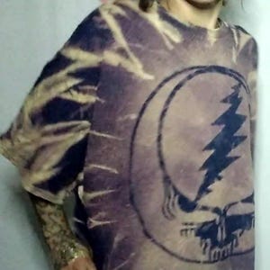 May include: Tie-dye t-shirt with a Steal Your Face graphic. The shirt features a purple and navy blue pattern. The graphic is a skull with a lightning bolt. The sleeves are long and the shirt is loose fitting.