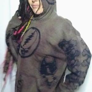 Pode incluir: Um moletom com capuz marrom com desenhos gráficos pretos. O moletom apresenta um design de raio, um design de morcego e desenhos florais nas mangas. A pessoa está usando calças rosa.