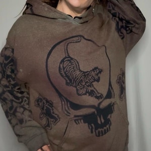 May include: A brown hooded sweatshirt with a black and white graphic print of a tiger inside a skull. The graphic is reminiscent of the Grateful Dead's iconic Steal Your Face logo.