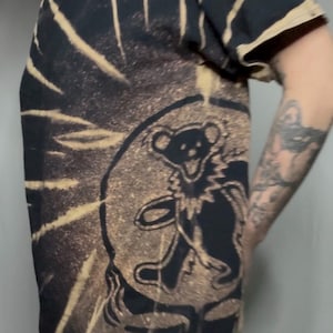 May include: Black t-shirt with a tie-dye design and a graphic of a dancing bear over a skull. The shirt has a V-neck and short sleeves. The bear and skull are in black, with a tan tie-dye pattern.