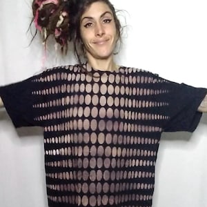 May include: Black short-sleeved shirt with a pattern of evenly spaced, light-colored ovals. The shirt has a crew neck and a loose fit. The design covers the front and sleeves of the shirt. The shirt is worn by a person.