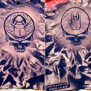 May include: A tie-dye t-shirt with a dark blue design on a light pink and purple background. The front features a skull with a scarab beetle design, and the back has a skull with a hand symbol. The text "JENZIFIED DESIGNZ" is at the bottom.