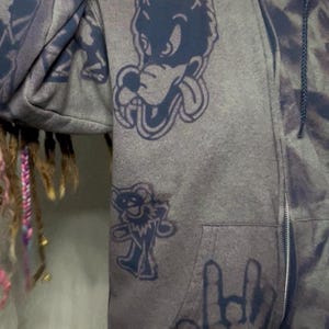 May include: Gray zip-up hoodie with dark blue graphics. The hoodie features a cartoon wolf, a bear, and a hand making a rock and roll gesture. The fabric has a tie-dye effect. The hoodie has a front pocket and a zipper closure.