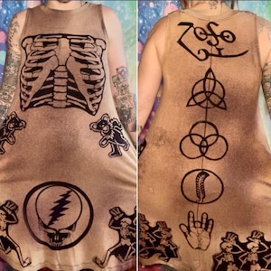 Puede incluir: Vestido sin mangas de color beige con diseños gráficos negros. La parte delantera muestra una caja torácica, osos bailarines y una calavera con un rayo. La parte trasera tiene la palabra "Zoso" y otros símbolos. El vestido tiene un aspecto vintage descolorido.