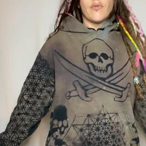 May include: A gray hoodie with a black skull and crossbones design, a geometric pattern on the sleeve, and a black geometric pattern on the front. The hoodie is worn by a person with long, colorful dreadlocks.