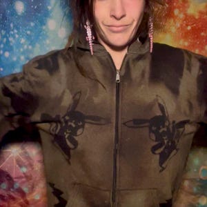 Pode incluir: Um casaco com capuz e zíper com padrão de camuflagem verde-oliva e castanho, com desenhos gráficos pretos de coelhos e raios. O zíper é prateado.