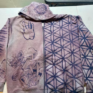 May include: A zip-up hoodie in a muted purple hue, adorned with dark blue botanical and geometric designs. The left side features a fern, butterflies, mushrooms, and a hand symbol. The right side showcases a symmetrical geometric pattern. The hood has a sunflower design.