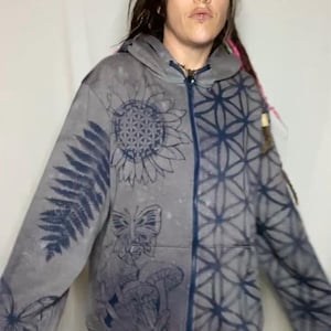 May include: A gray zip-up hoodie with a blue and white geometric pattern and a sunflower design. The hoodie has a fern design on one sleeve and a butterfly design on the other sleeve.